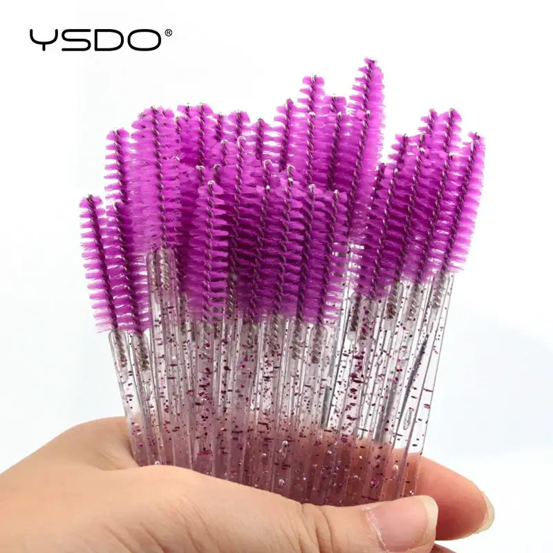 Hand holding a bunch of disposable eyelash spoolie brushes with bright purple bristles and clear, glittery handles.