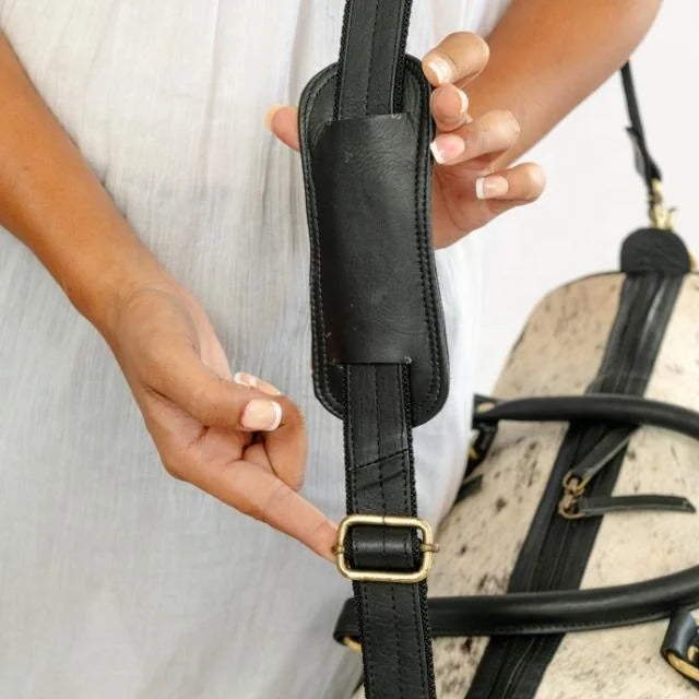 Unisex cow hair-on-hide weekender duffel bag with black leather shoulder strap and adjustable brass hardware.