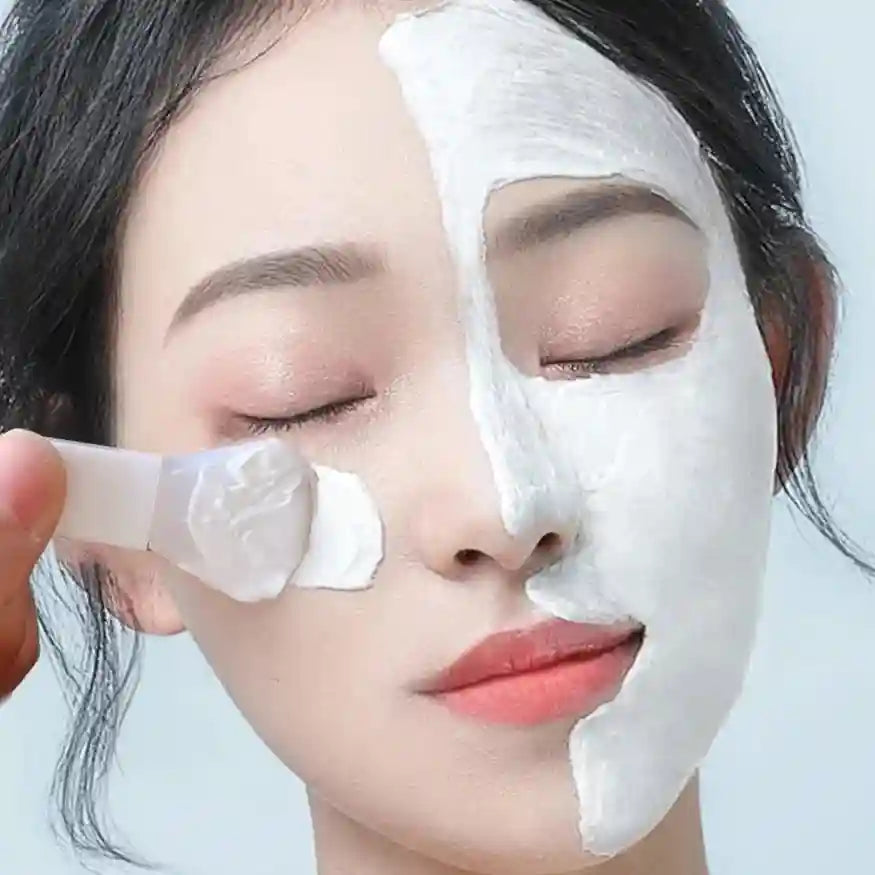 A hand uses a white silicone applicator brush to apply a thick, white mud mask to a woman's face.