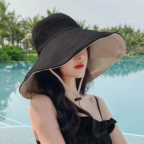 Tidesmit reversible wide-brim sun hat in black and beige with a chin strap, worn by a woman by a swimming pool.