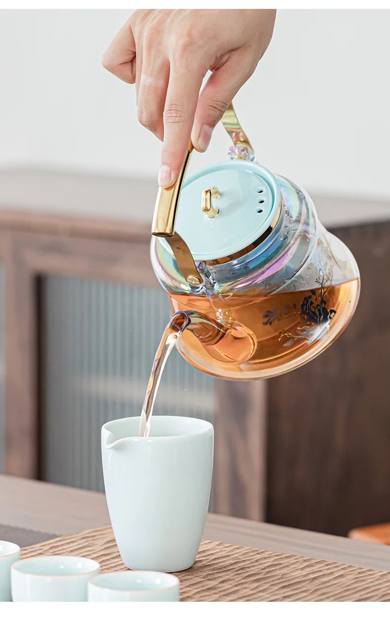 Hand pouring tea from an iridescent glass teapot with a light blue lid and gold handle into a ceramic cup.