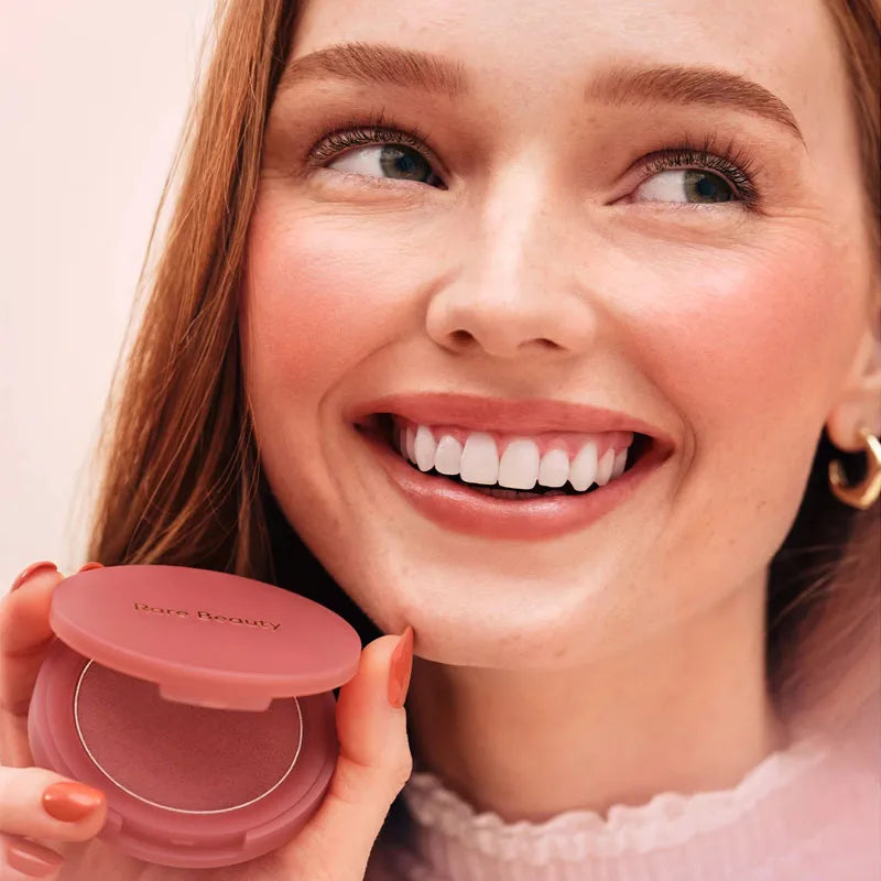 Model holding a Rare Beauty powder blush compact with smooth, matte pink pigment and soft-focus finish.