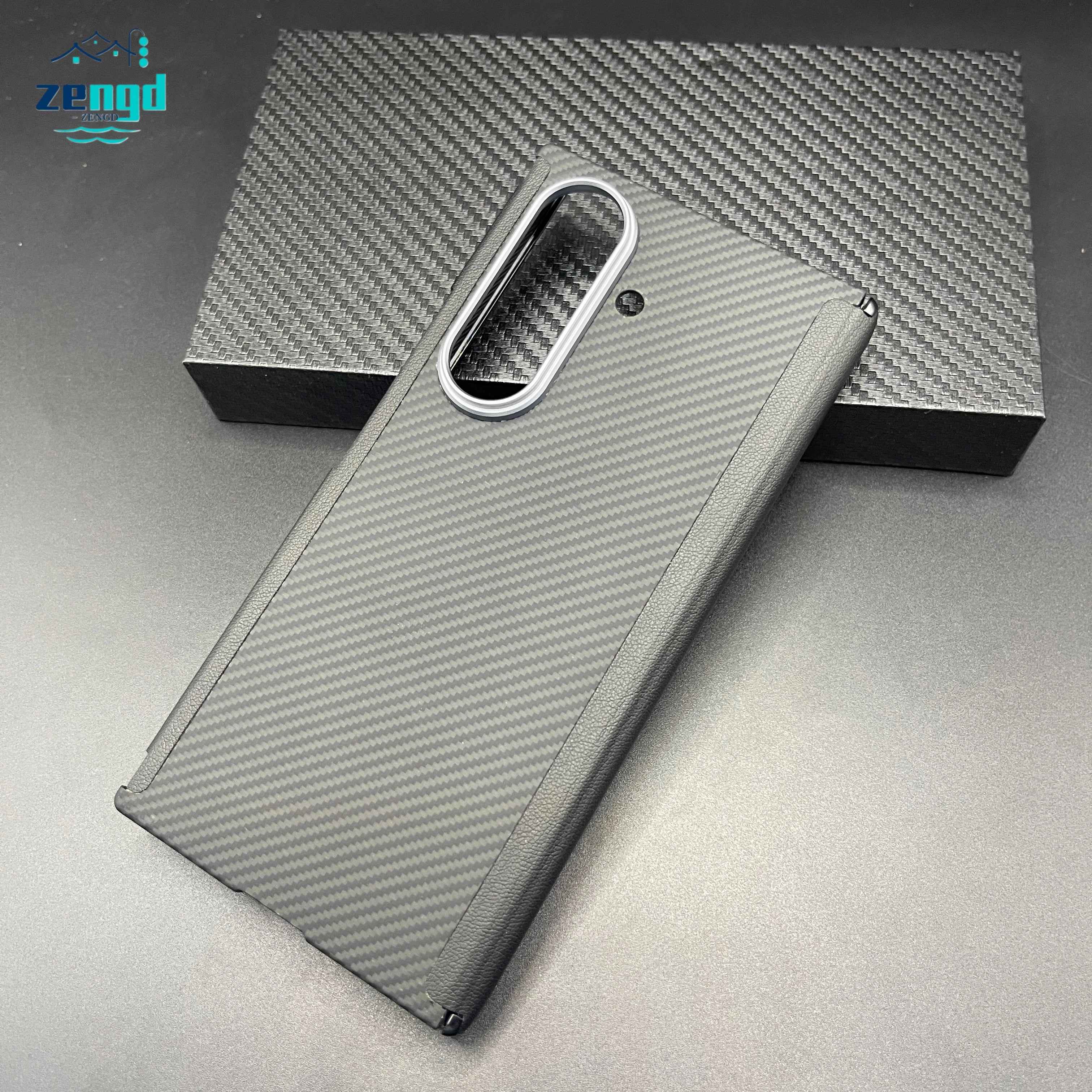Black carbon fiber phone case with a silver camera cutout for a foldable smartphone, resting on a matching box.