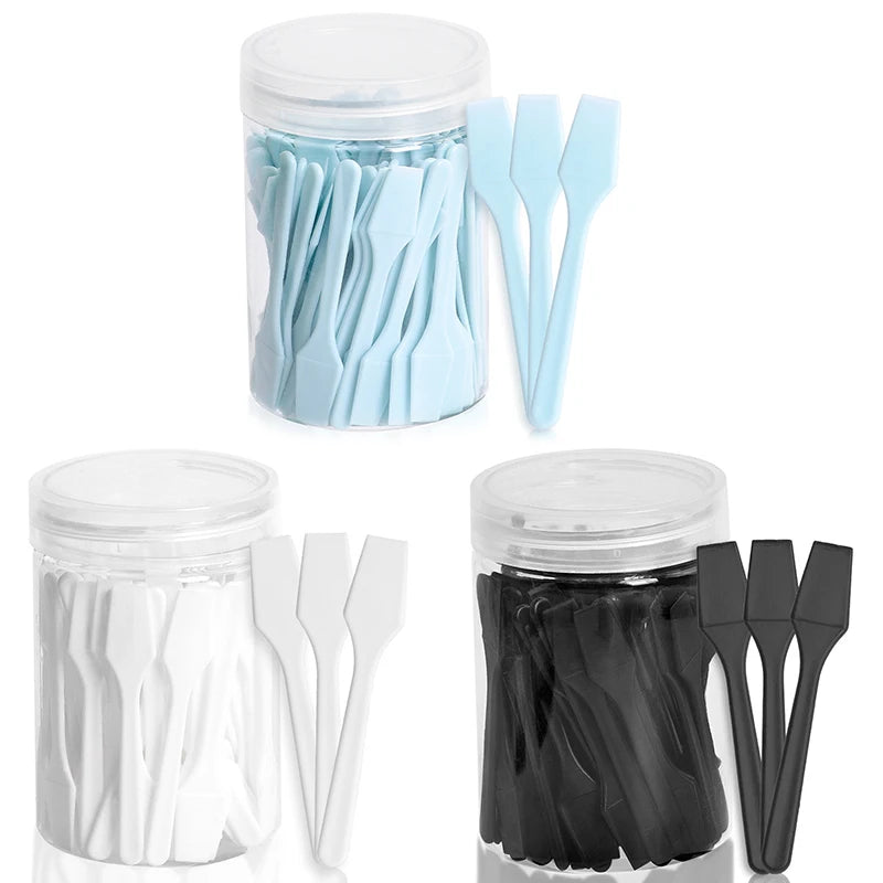 Three clear plastic jars filled with small, flat cosmetic spatulas in light blue, white, and black colors.