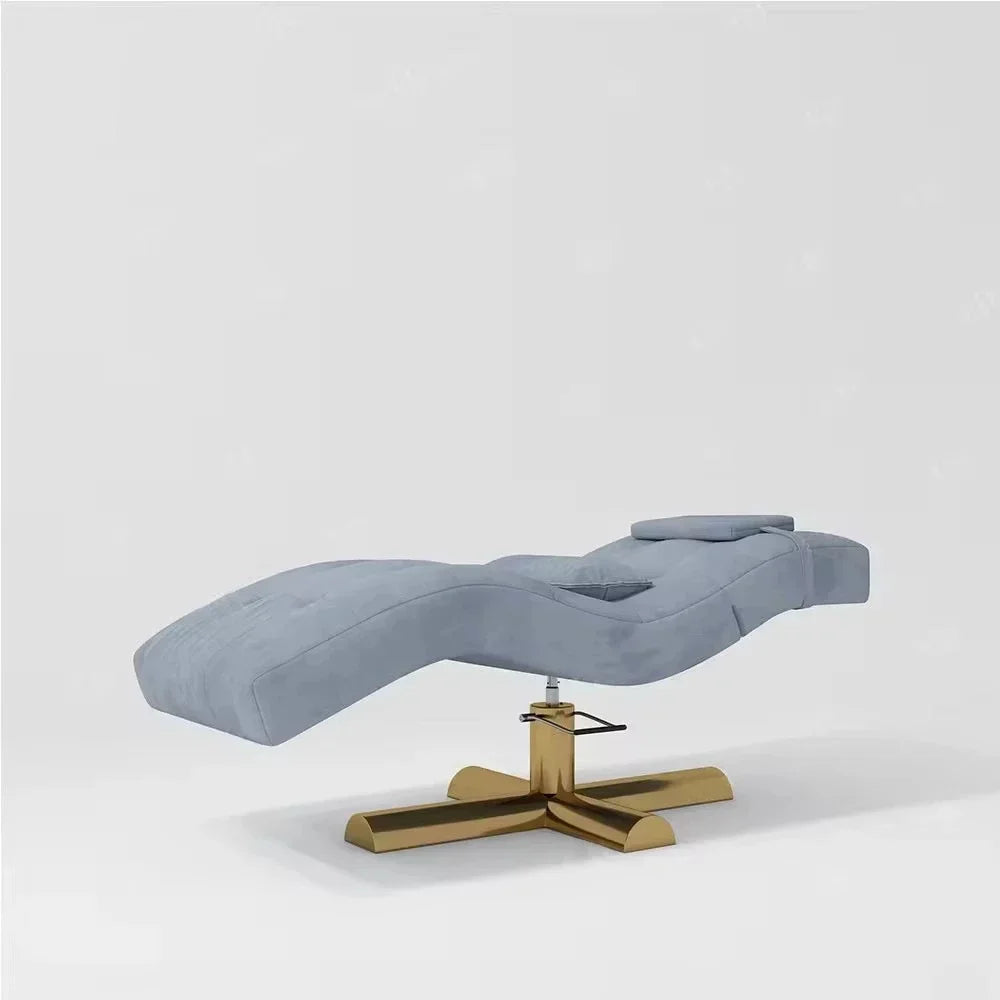 Light blue velvet facial treatment bed with adjustable headrest on a gold-finished, four-pronged metal pedestal base.