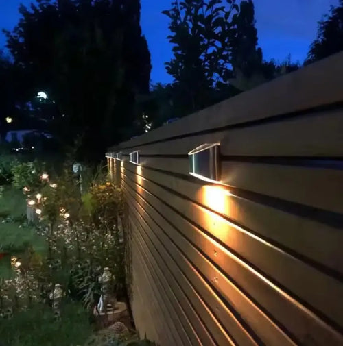 Stainless steel solar outdoor wall lights mounted on a wooden fence, casting warm light down.