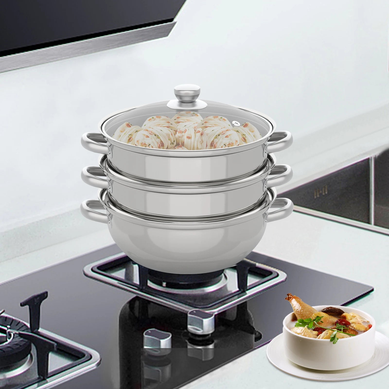 Stacked stainless steel steamer with clear lid on cooktop, curved side handles.