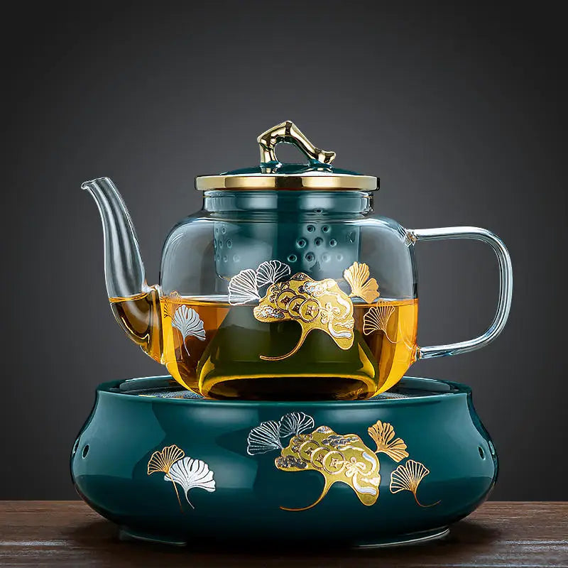 Glass teapot with gold ginkgo leaf pattern on a matching dark green ceramic warming base.
