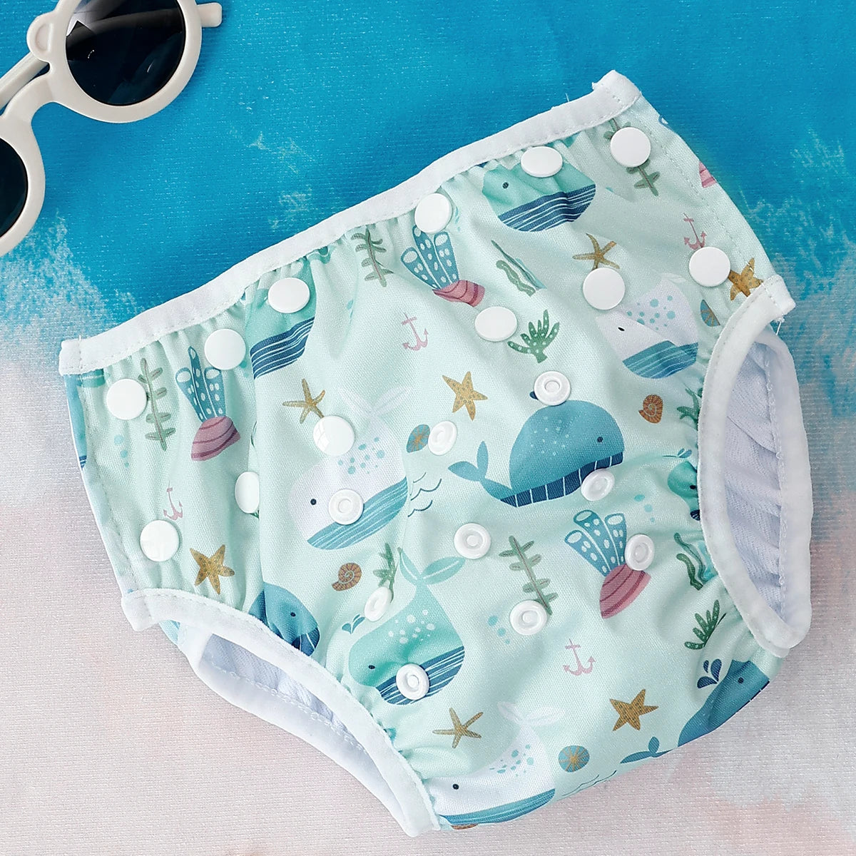 Reusable cloth baby diaper with adjustable snaps and cute whale and sea life print on a light blue background.