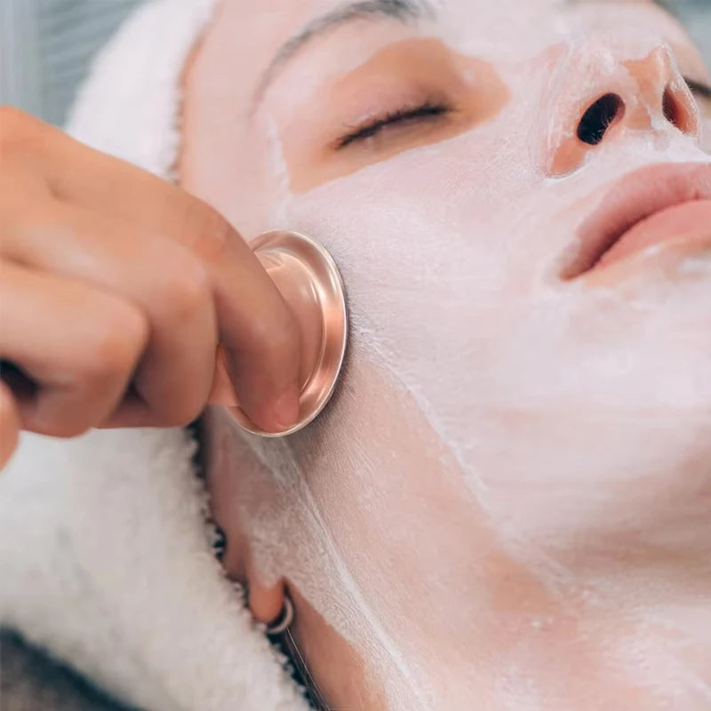 Hand using a metallic rose-gold silicone facial cupping tool on a person with a white face mask.