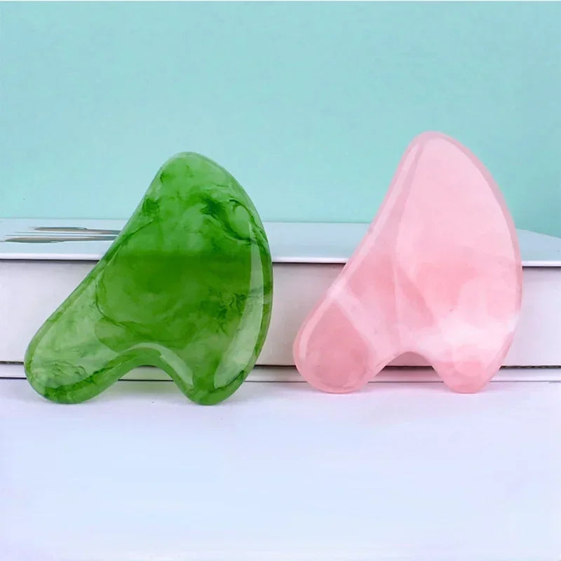 Two smooth, heart-shaped resin gua sha massage tools, one marbled green and one translucent pink, against a blue wall.