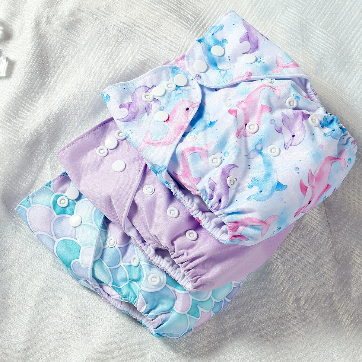 Set of 3 reusable baby cloth diapers with dolphin and mermaid scale prints, adjustable snap buttons.