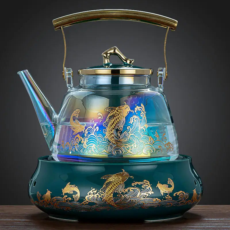 Iridescent glass teapot with gold koi fish pattern on a teal ceramic warming base, featuring a brass swing handle.