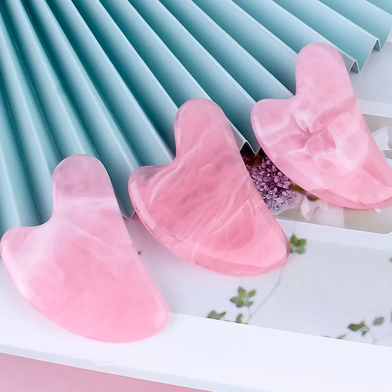 Three heart-shaped, pink marbled resin gua sha massage tools resting on a white surface with a blue pleated background.