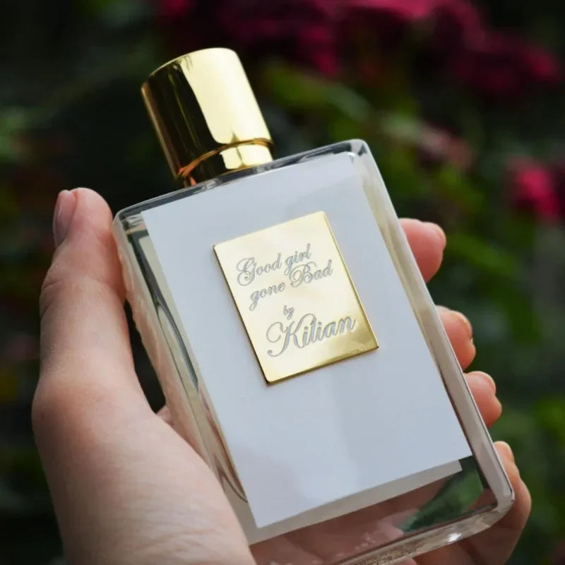 Kilian Good Girl Gone Bad 50ml perfume bottle with a gold cap and label held in hand against a blurred floral backdrop.