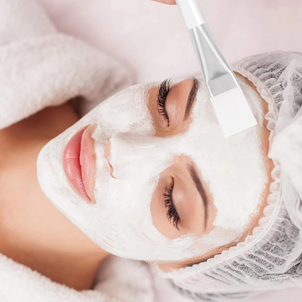 A flat, white facial mask applicator brush with a transparent handle applying a white mask to a woman's face.