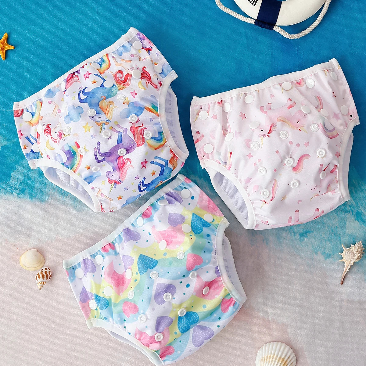Three reusable baby cloth diapers with unicorn and heart prints on a beach-themed background.