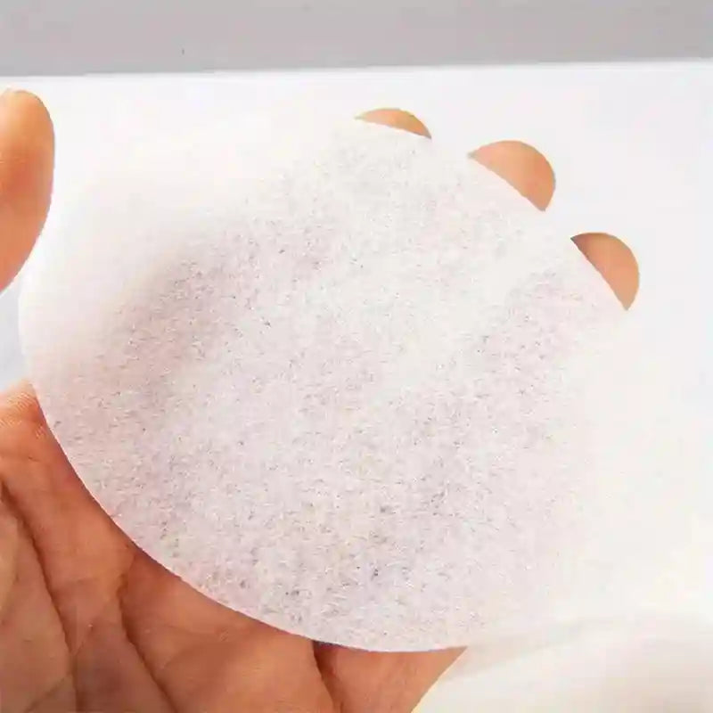 A hand holding a thin, round, white disposable cotton eye mask paper for DIY skincare.