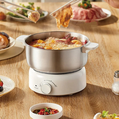 Stainless electric cooking pot with glass lid, white base, and bubbling hot soup inside