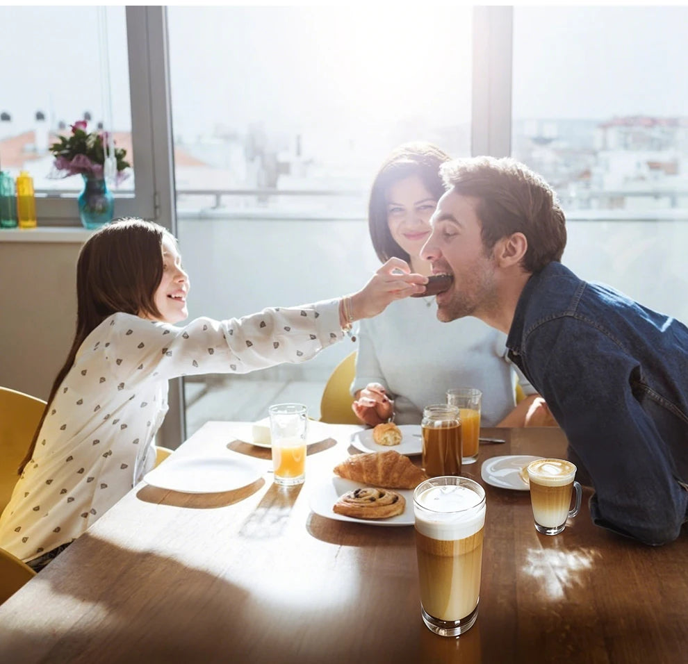 Family enjoying lattes in clear glass mugs on a wooden breakfast table in a sunlit dining room.