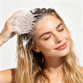 Woman using a white, silicone scalp massager brush with thick bristles on soapy, wet blonde hair.
