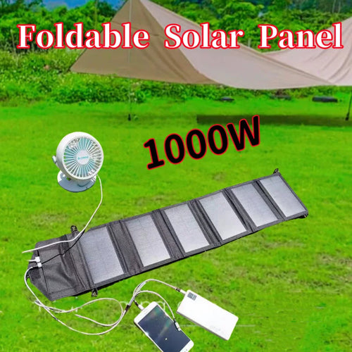 Foldable black solar panel on green grass, powering a white desk fan, mobile phone, and power bank via charging cables.