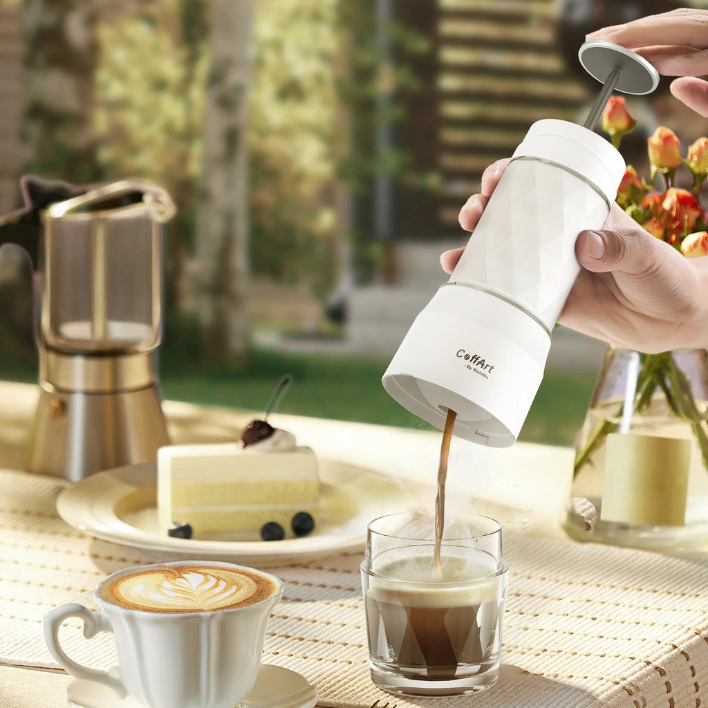 White CoffArt portable manual coffee maker pouring espresso into a glass on an outdoor table.