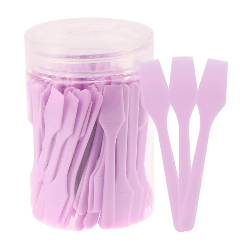 A clear plastic jar filled with 100 small, light purple plastic cosmetic spatulas for skincare application.