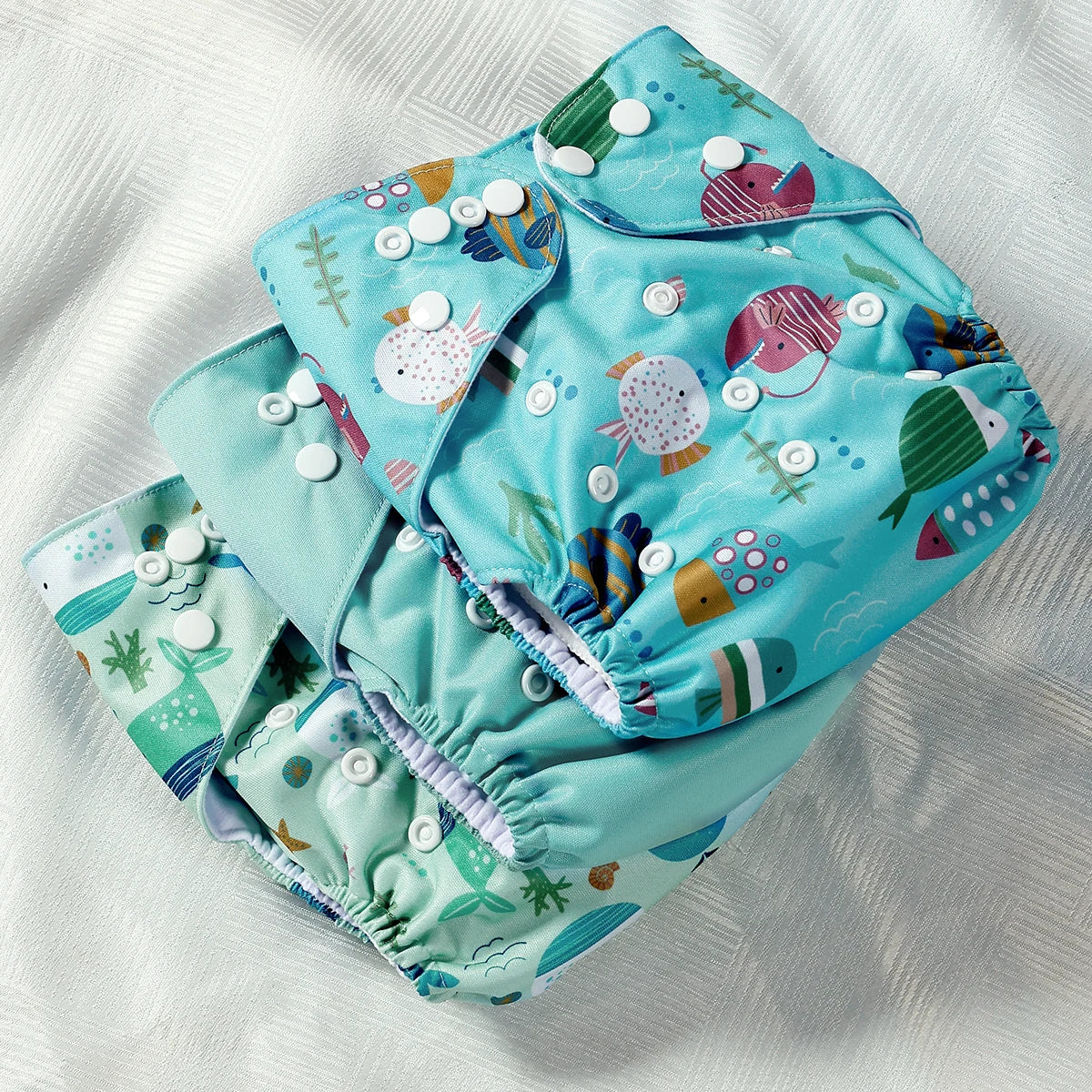 Reusable baby cloth diaper in teal with colorful fish patterns and adjustable snap closures.