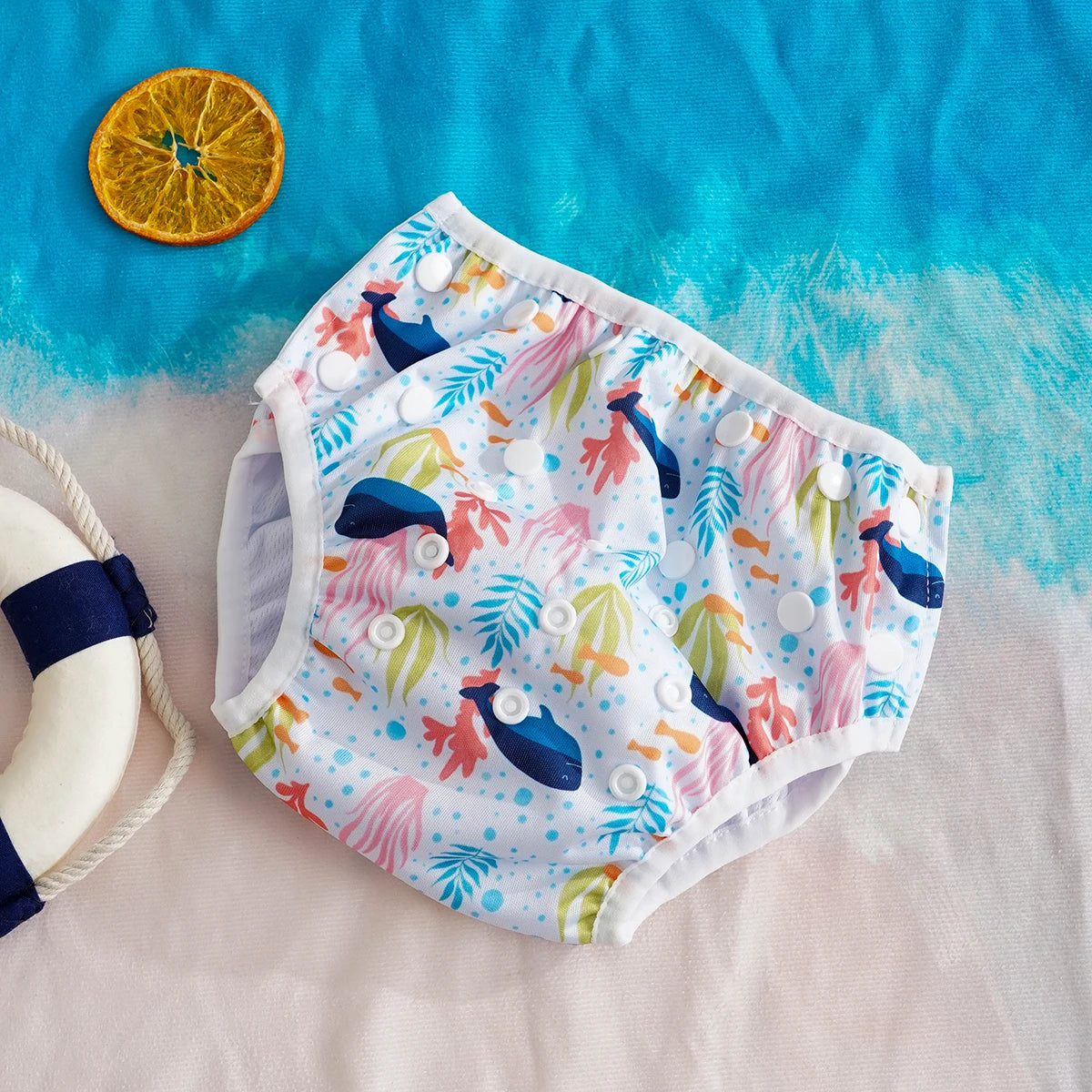 Reusable baby swim diaper with snap closures, featuring a colorful whale and coral ocean print on a beach background.