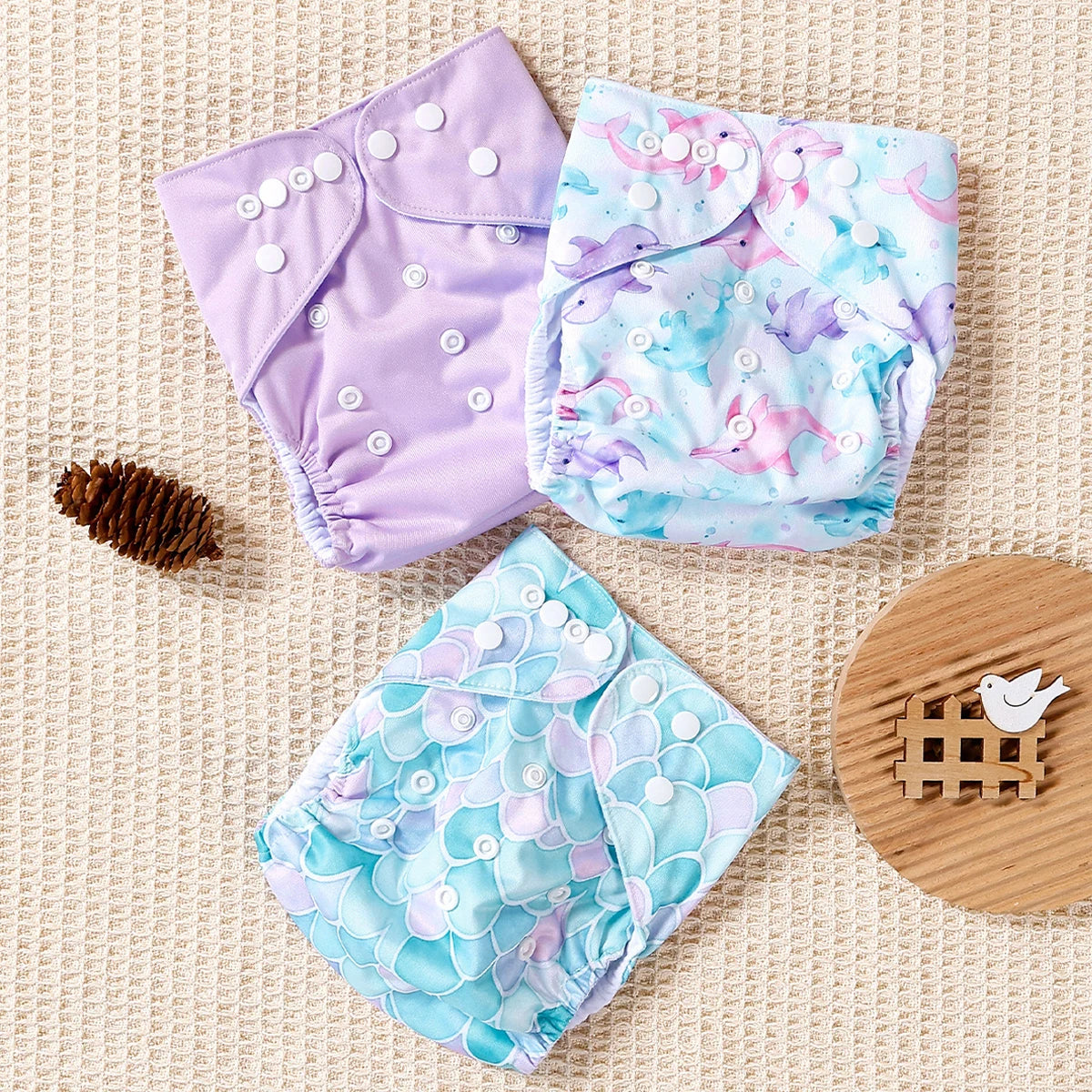 Three reusable cloth baby diapers featuring purple, dolphin, and mermaid scale patterns with adjustable snap closures.