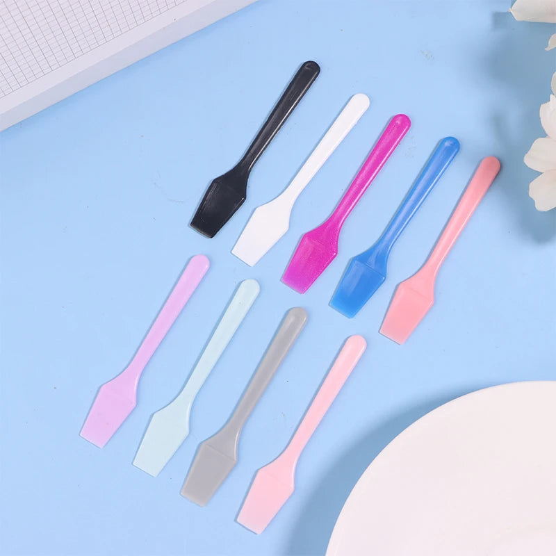 A set of small, colorful plastic cosmetic spatulas for applying creams and masks, arranged on a light blue background.